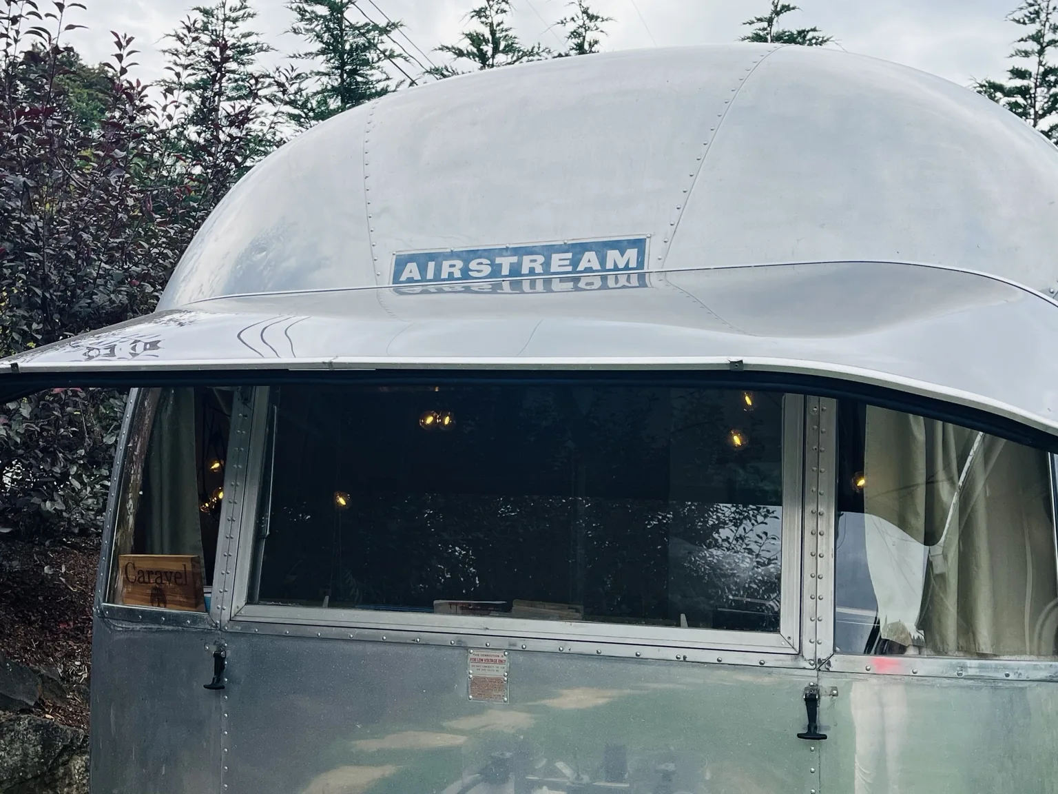 Actual Caravel Airstream exterior, showing its curved silver aluminum front and large window among trees